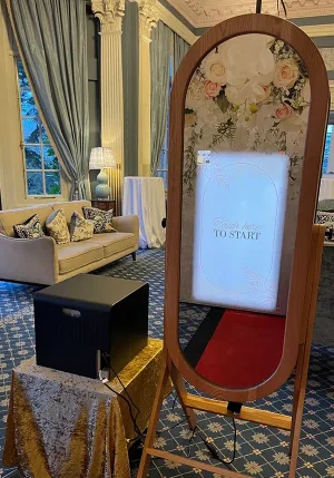 Elegant oval mirror photo booth setup at a luxury wedding venue with floral backdrop and red carpet.